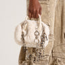 Close-up of a person holding the APRES SHERPA BAG by Rag Company LLC, a small white faux fur purse adorned with multiple silver chains and metallic charms. The person is wearing light-colored cargo-style pants with several large pockets. The background is a plain, light shade.