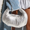 A person wearing a textured blue garment holds a large, puffy PUFFER DREAMS PURSE in front of their hip. The outfit also features a fringed blue covering, metallic embellishments, and a removable silver link chain strap. The neutral background emphasizes the striking fashion pieces.