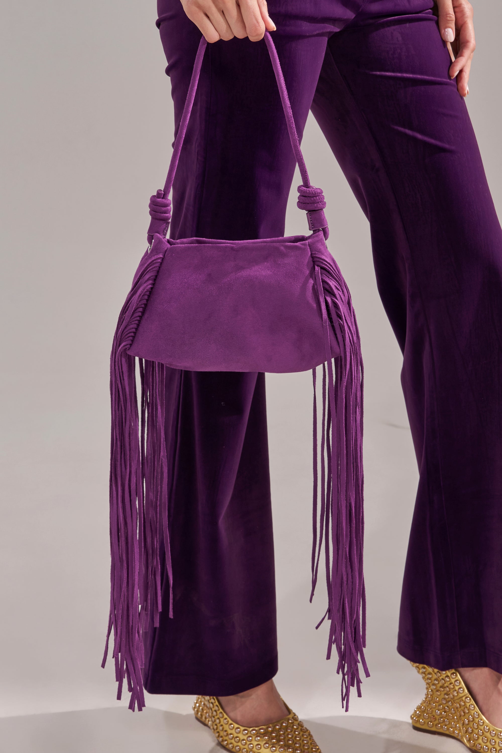 A person in purple pants holds the IN THE WIND SUEDE PURSE, featuring long fringe. Gold-studded shoes peek out, set against a neutral background.