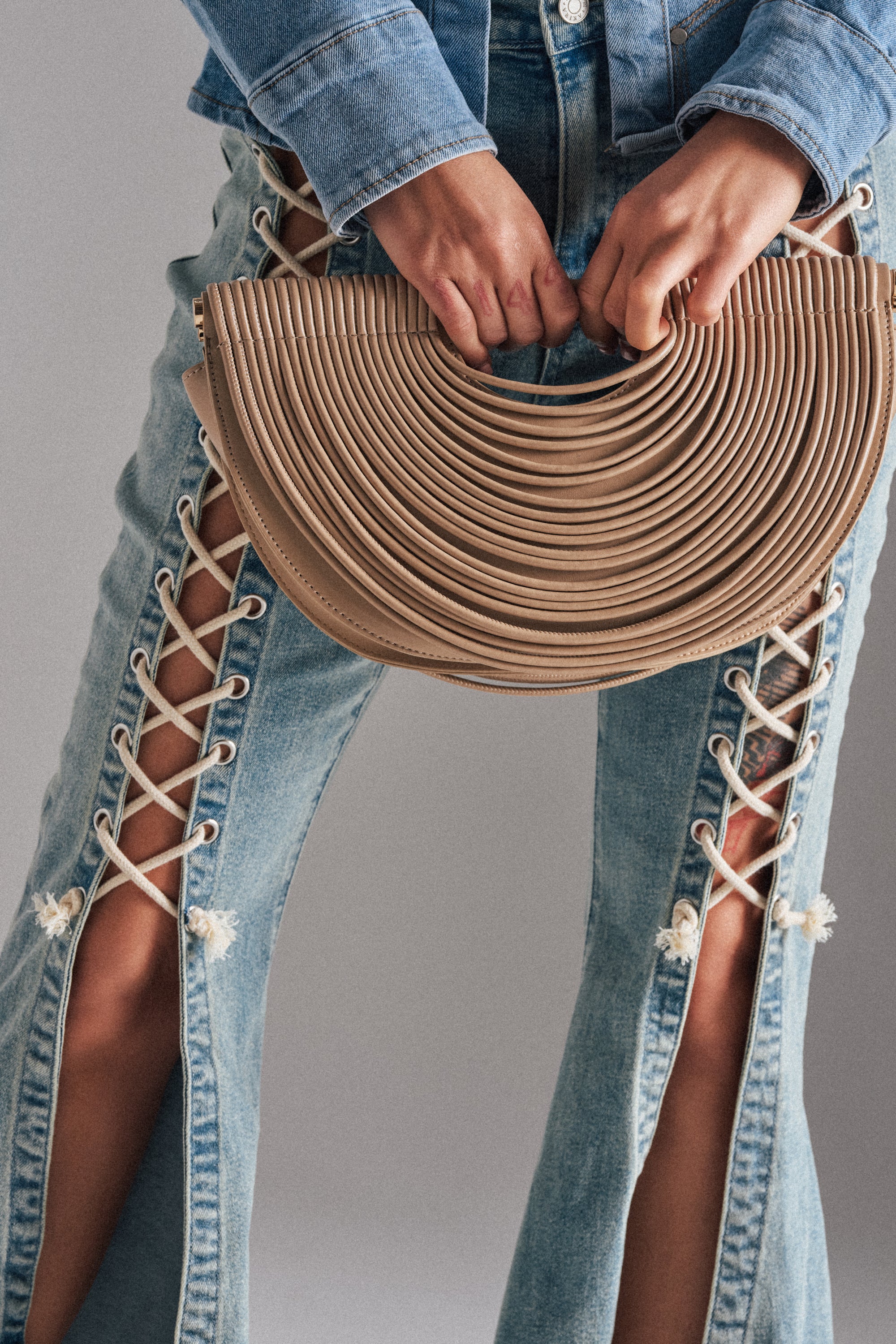 Wearing lace-up denim jeans and a denim jacket, a person holds the CRESCENT CARRY-ALL PURSE—featuring a half-moon silhouette and ribbed texture—in both hands in front of their legs.
