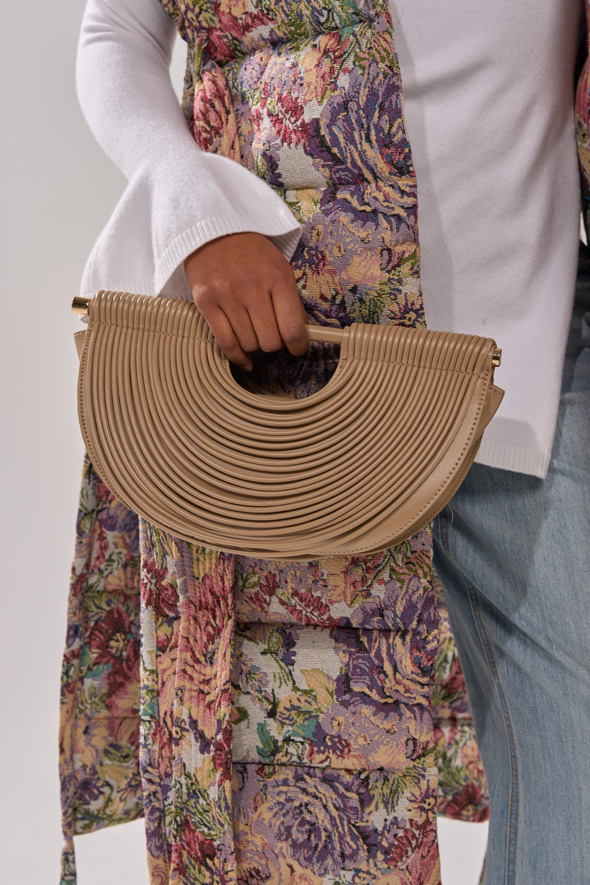 A person in a floral-patterned coat, white top, and blue jeans holds the CRESCENT CARRY-ALL PURSE—a beige faux leather handbag with a textured finish, curved handle, and half-moon silhouette.
