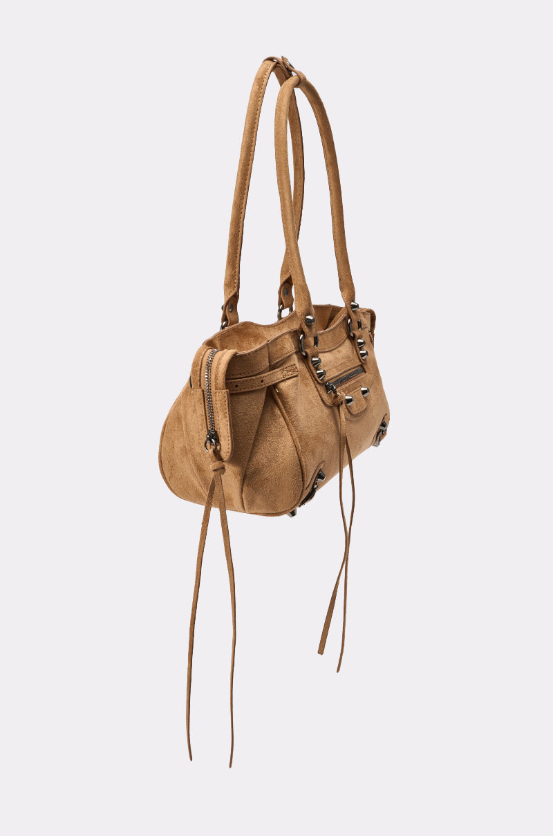The IN THE CITY BAG is a tan brown suede shoulder bag with double handles, silver hardware, decorative studs, and long leather tassels on the zipper and sides—ideal for streetwear styling. Photographed on a light plain background. 2