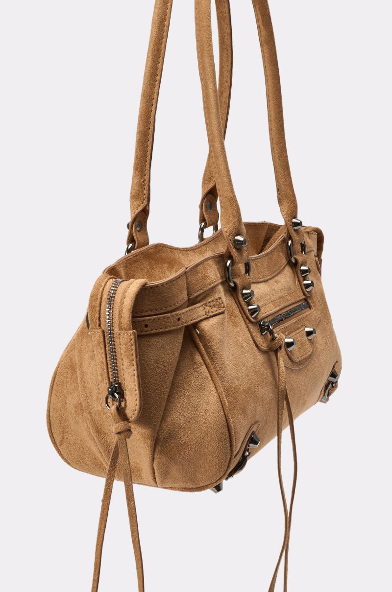 The IN THE CITY BAG is a brown suede shoulder bag featuring dual handles, silver hardware, a front zipper pocket, and decorative tassels, photographed against a plain light background. 3