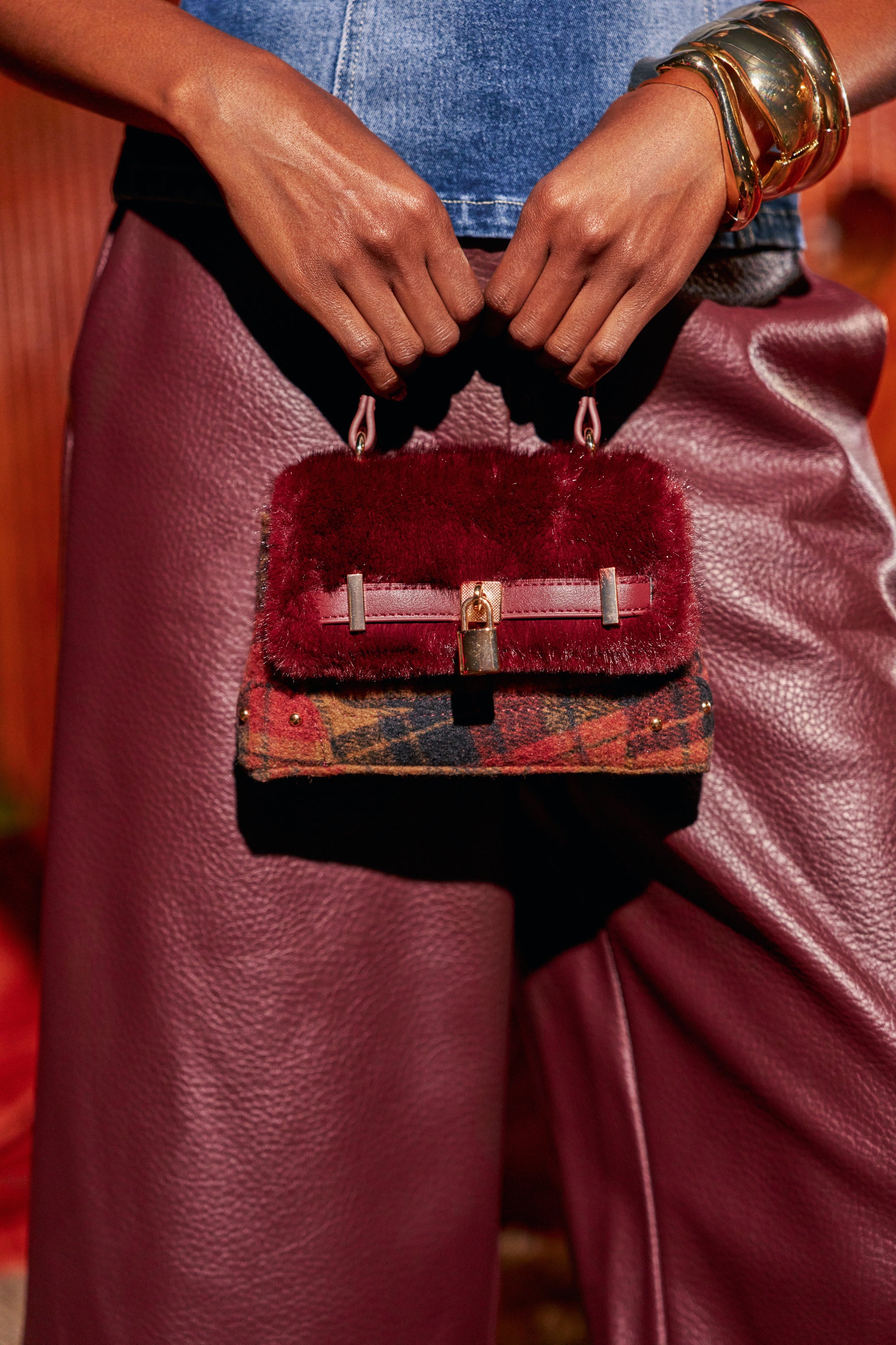 A person holds a LOCKED IN MINI PURSE, a small, structured plaid flannel bag featuring a plush, deep red top flap with a gold-toned padlock closure.