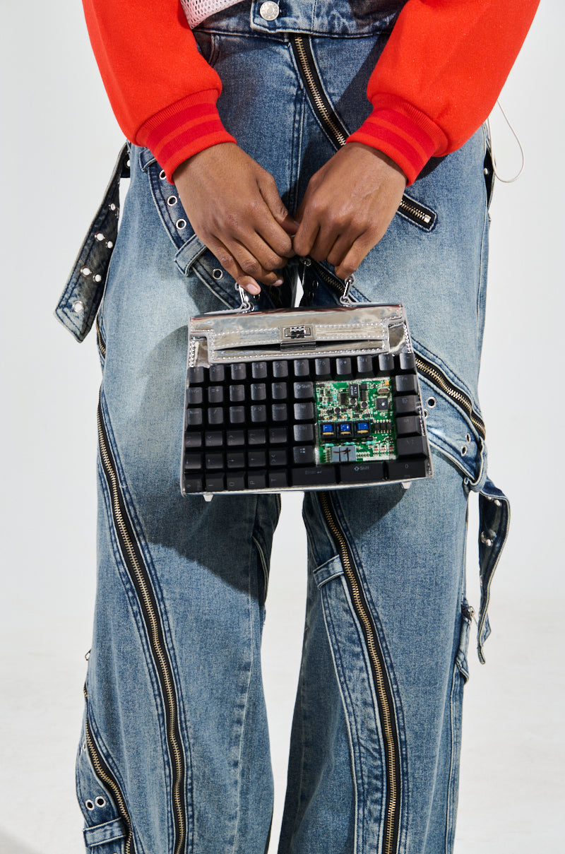 A person holds the TYPE IT OUT KEYBOARD PURSE, crafted from a recycled computer keyboard and circuit board. They wear denim jeans with multiple zippers and a bright red shirt, perfectly complementing this innovative accessory.