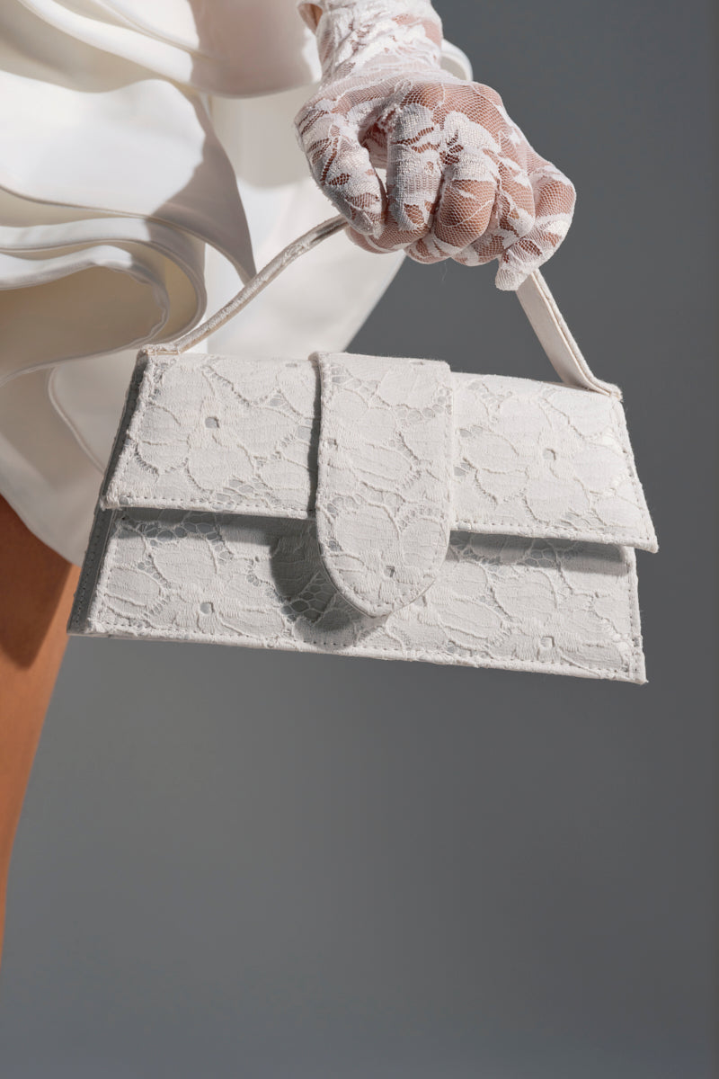 A person in sheer white lace gloves holds the HONEYMOON LACE APPLIQUE BAG, featuring a delicate floral lace overlay. The white dress, bare leg, and gray background highlight the bag’s elegant and sophisticated appeal.