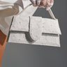 A person in sheer white lace gloves holds the HONEYMOON LACE APPLIQUE BAG, featuring a delicate floral lace overlay. The white dress, bare leg, and gray background highlight the bag’s elegant and sophisticated appeal.
