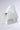 The HONEYMOON LACE APPLIQUE BAG is a chic white handbag with textured lace and an elegant top handle. It has a curved flap closure and a detachable chain strap for versatility, set against a neutral gray background.