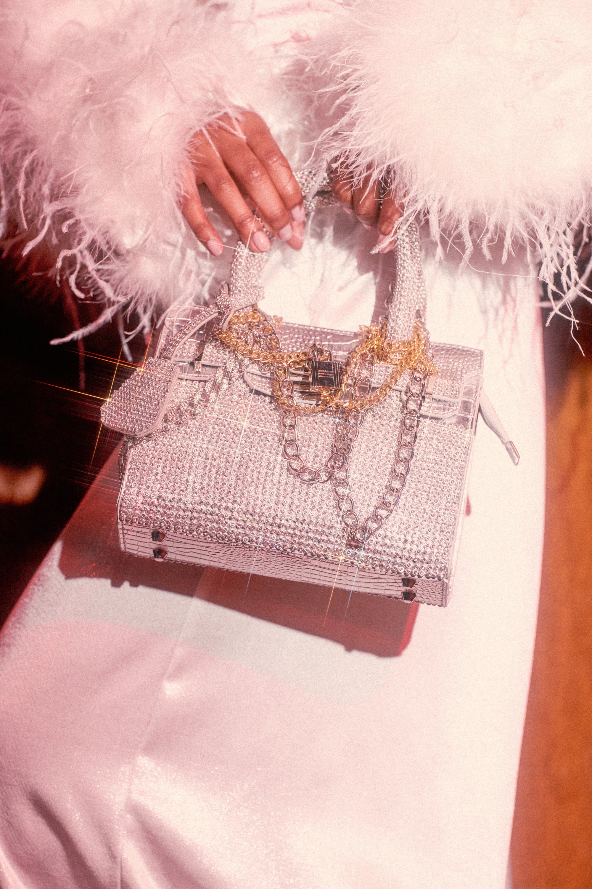 A person in a white, feather-trimmed outfit holds the TOP NOTCH EMBELLISHED CHAIN PURSE, adorned with jewels and chains, while soft lighting casts a glamorous, vintage effect.