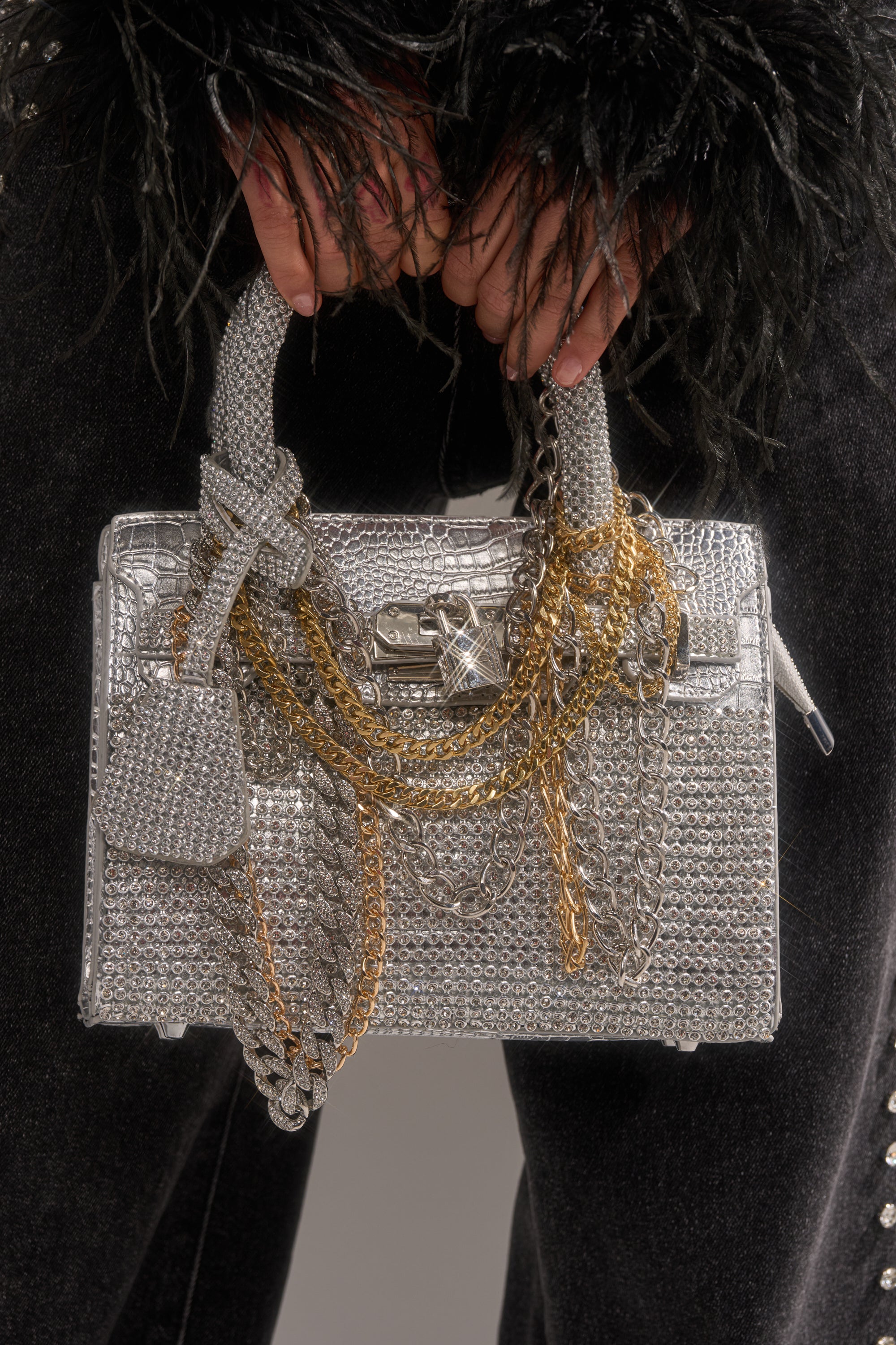 A person in black, textured sleeves holds the TOP NOTCH EMBELLISHED CHAIN PURSE—a metallic silver rhinestone handbag with layered gold and silver chains, a chic clasp, and a matching charm for extra style.