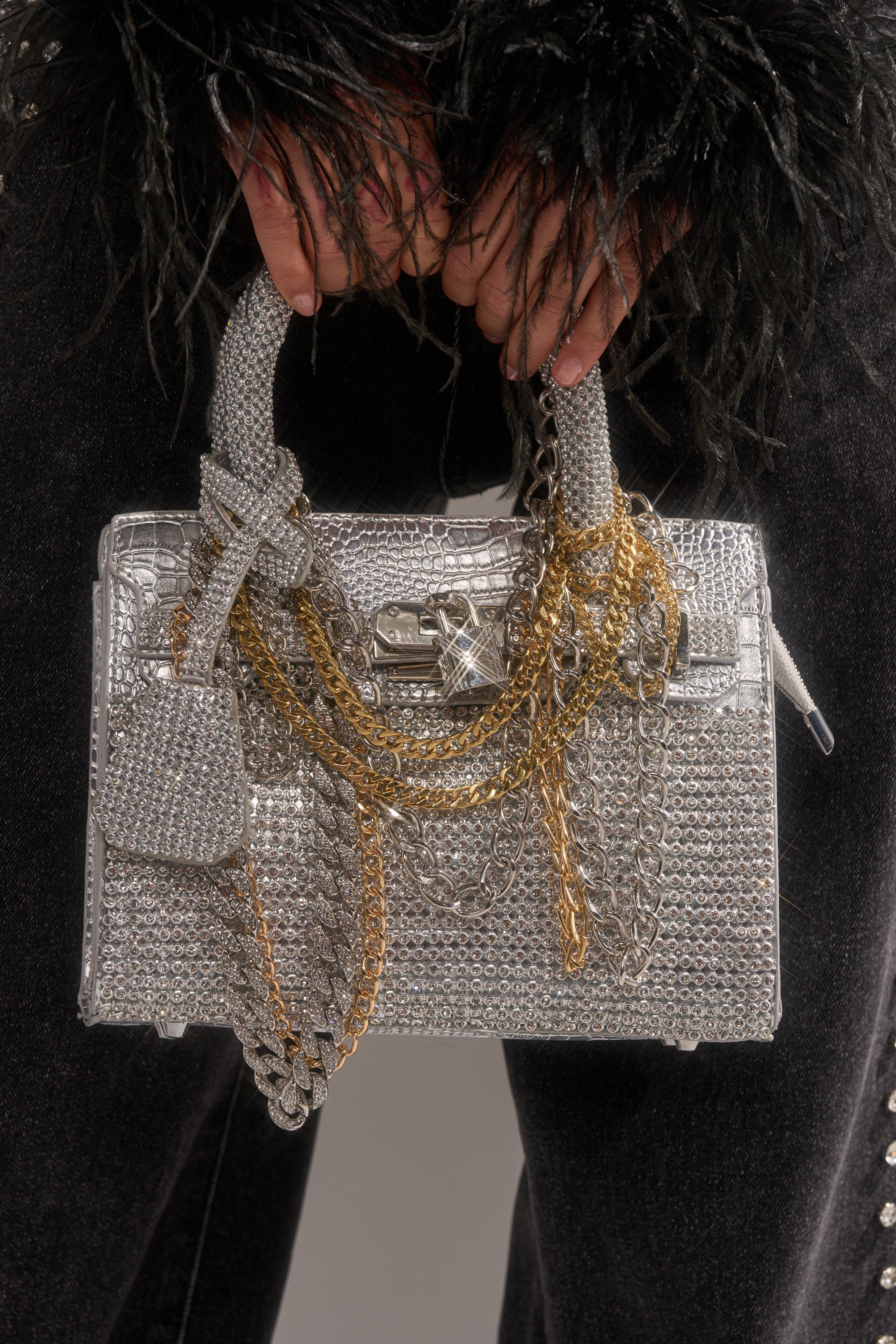 A person in black, textured sleeves holds the TOP NOTCH EMBELLISHED CHAIN PURSE—a metallic silver rhinestone handbag with layered gold and silver chains, a chic clasp, and a matching charm for extra style. 2