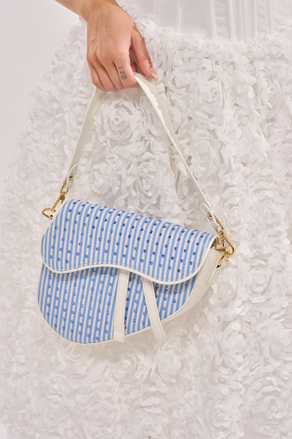 A person in a white, textured floral skirt holds the GONE SAILING STRIPE BAG, featuring gold hardware and a cream strap. 1