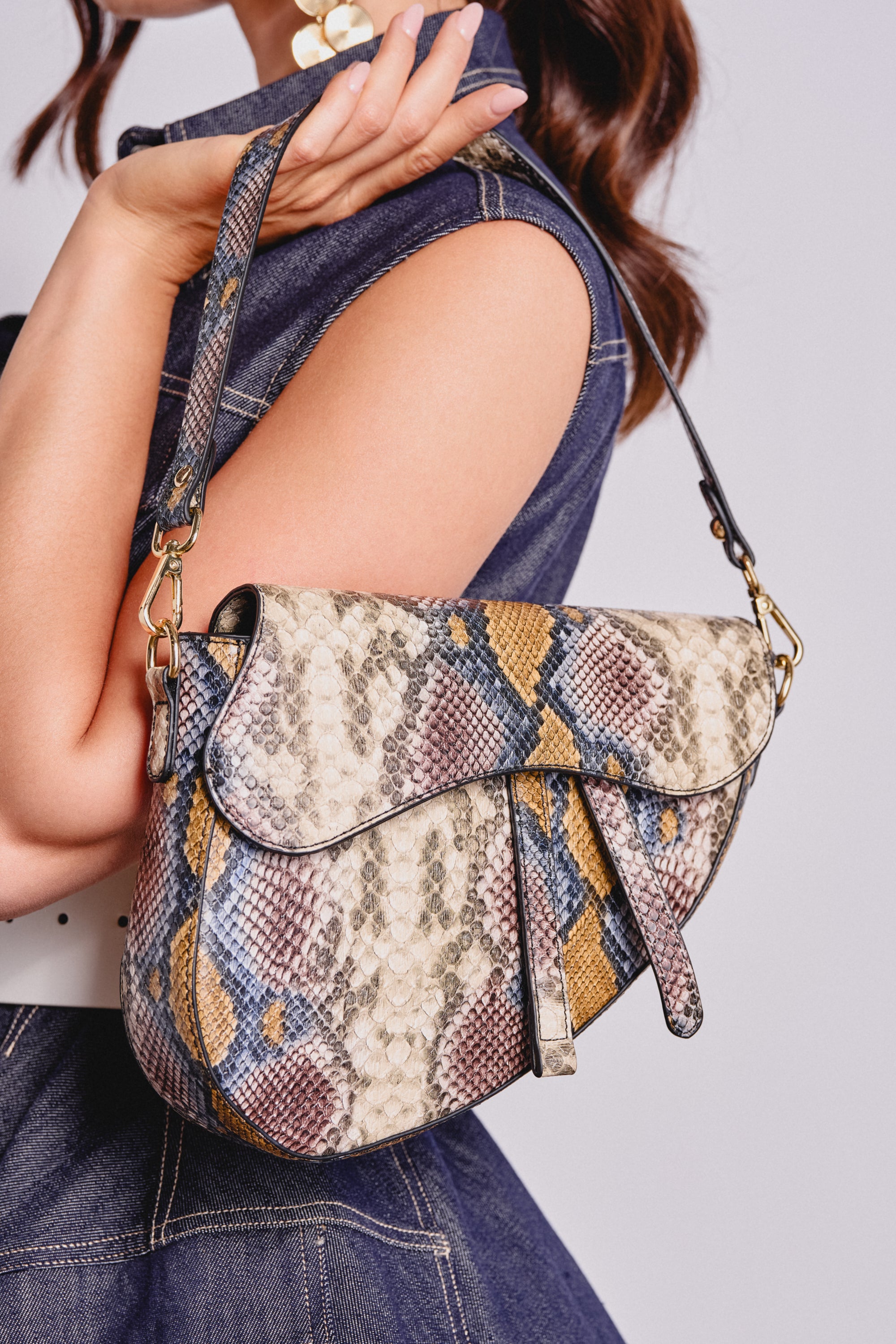 A woman holds the DO U GET IT PURSE, a small faux leather snakeskin bag in beige, blue, and yellow. She wears a sleeveless denim dress against a light purple background. 1