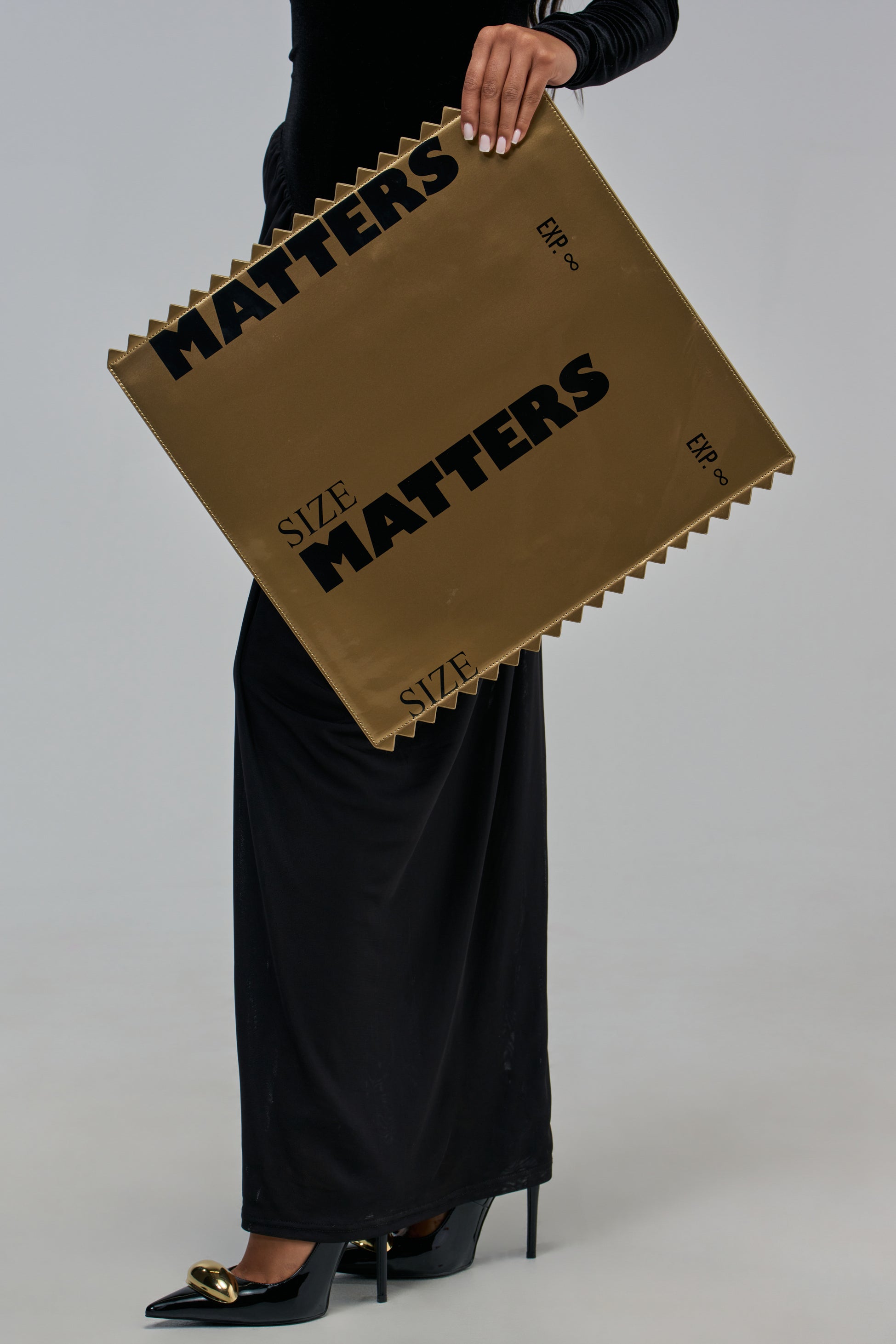 A person holding a SIZE MATTERS CHROME PURSE, an oversized gold clutch with serrated edges printed with the text “SIZE MATTERS” and “EXP. ∞”.