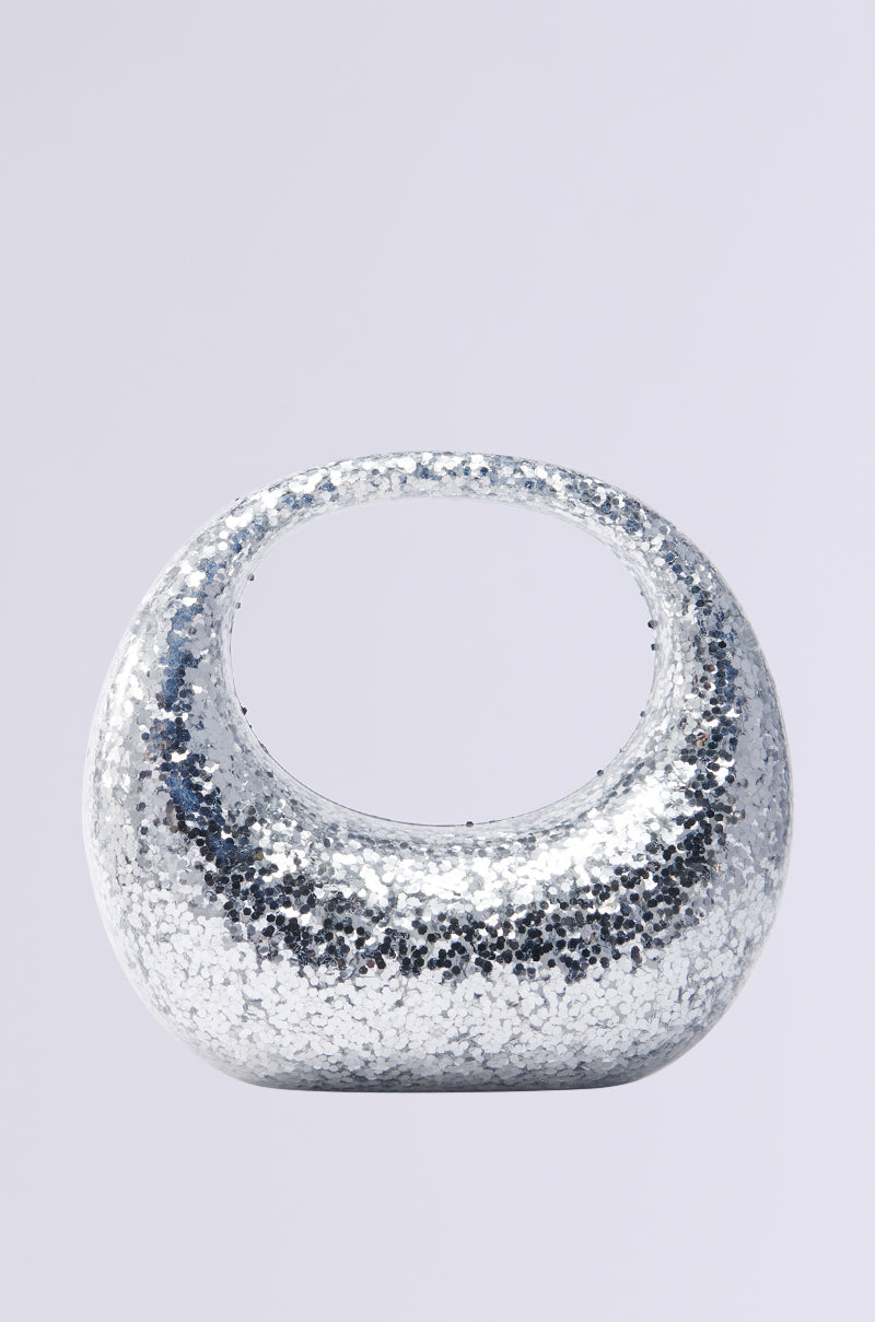 The ALWAYS ADD SPARKLE HARD SHELL PURSE is a glittering silver handbag with metallic sequin detailing, a rounded shape, and a wide circular handle, set against a light gray backdrop. 0