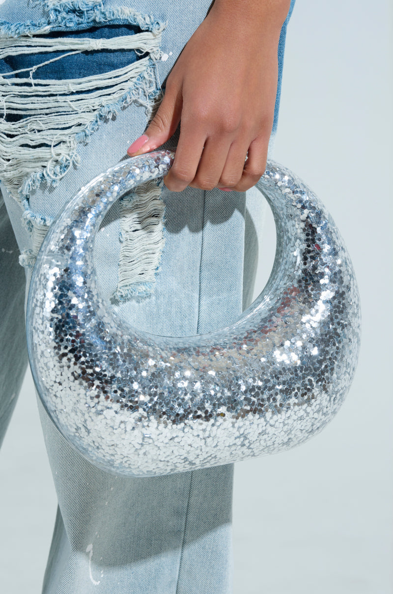 A person holds the ALWAYS ADD SPARKLE HARD SHELL PURSE, a sparkling crescent-shaped silver handbag with metallic sequin detailing. They're in light blue ripped jeans against a bright, minimalistic background that enhances the bag's chic allure. 1