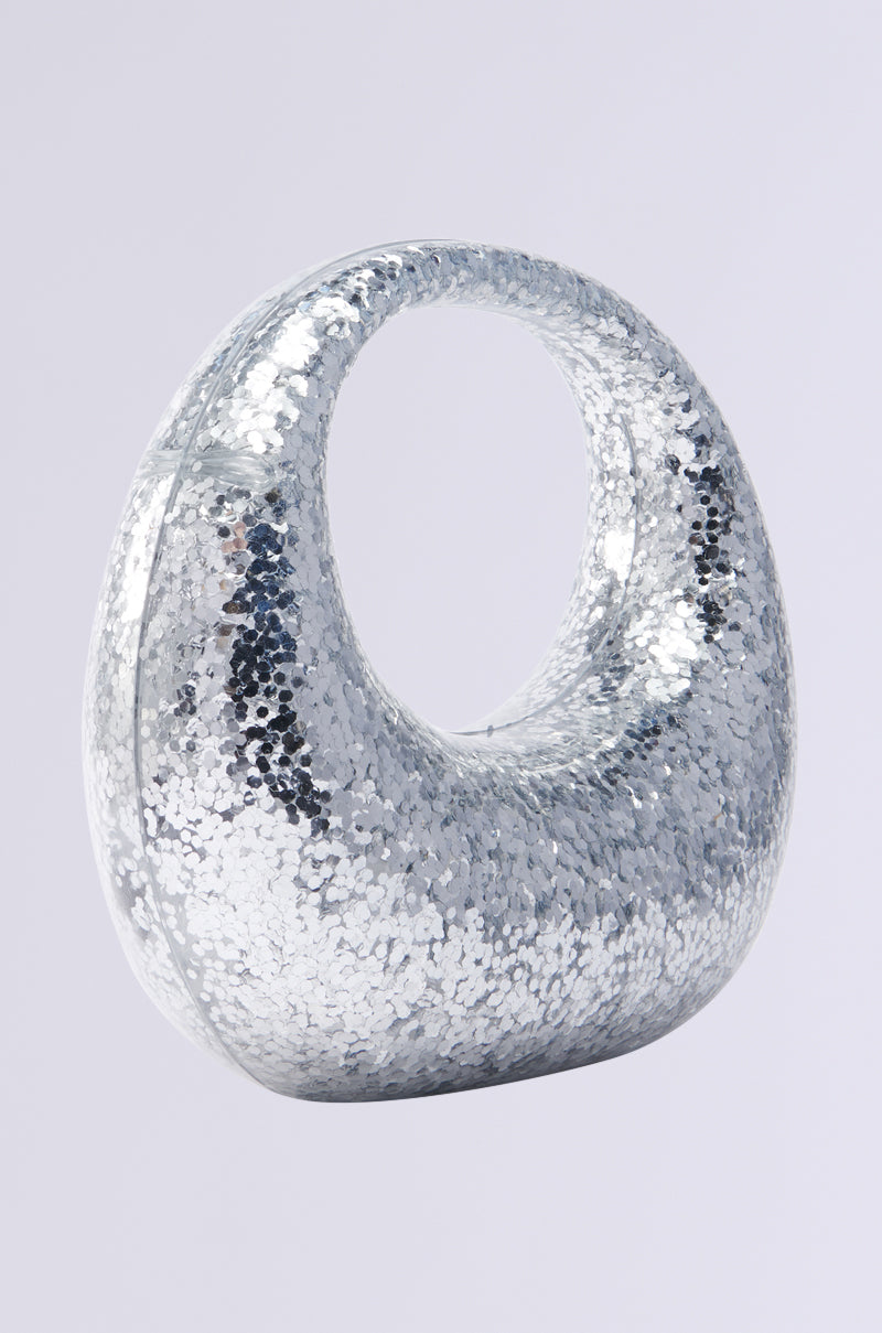 The ALWAYS ADD SPARKLE HARD SHELL PURSE is a shiny, silver handbag with metallic sequins and a unique curved shape, featuring a circular handle on top against a plain light gray background. 2