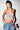 A woman poses against a white wall in the FLY WITH ME BUTTERFLY TOP, a vibrant sequin piece with adjustable straps, paired with low-rise jeans with metal chains. She accessorizes with long earrings, sleek hair, and a pink beaded bear purse.