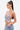 A woman with long, straight hair in a high ponytail wears large hoop earrings, layered necklaces, and the FLY WITH ME BUTTERFLY TOP—featuring sequins and adjustable straps—with light blue chain-detailed jeans on a white background.