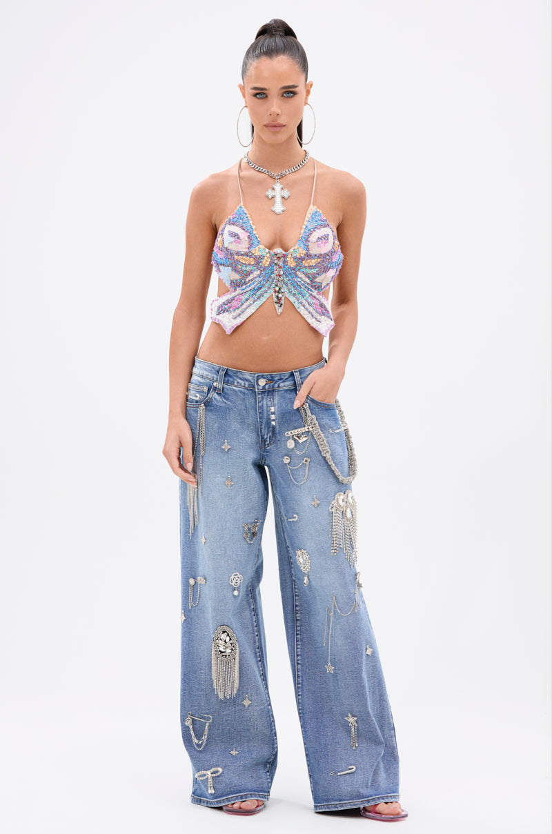 A woman with a high ponytail and confident expression poses against a white background in the sparkling FLY WITH ME BUTTERFLY TOP, wide-leg blue jeans with metallic charms, hoop earrings, layered necklaces, and pink heels. 5