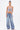 A woman with a high ponytail and confident expression poses against a white background in the sparkling FLY WITH ME BUTTERFLY TOP, wide-leg blue jeans with metallic charms, hoop earrings, layered necklaces, and pink heels.