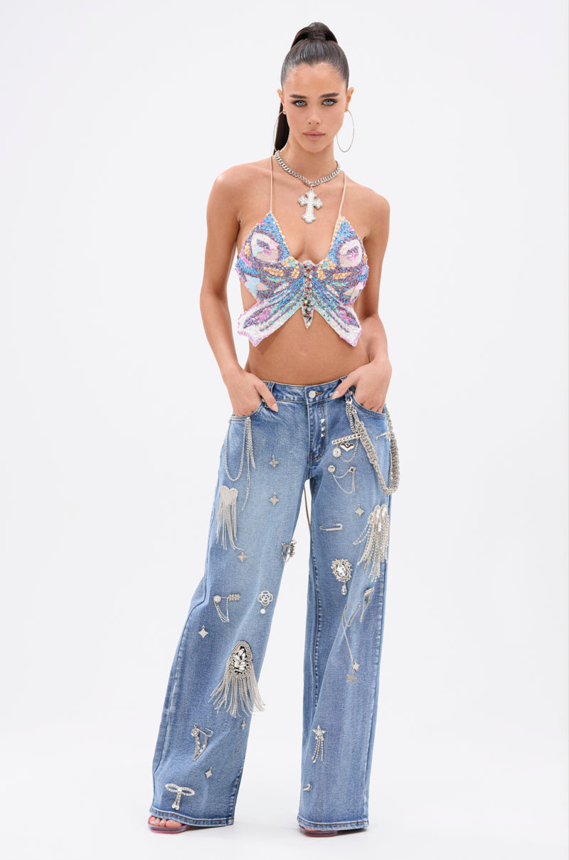 A woman poses against a white background wearing the FLY WITH ME BUTTERFLY TOP, featuring sequins and adjustable straps, paired with wide-leg jeans decorated with rhinestones, chains, and patches. She accessorizes with hoop earrings and a high ponytail. 6