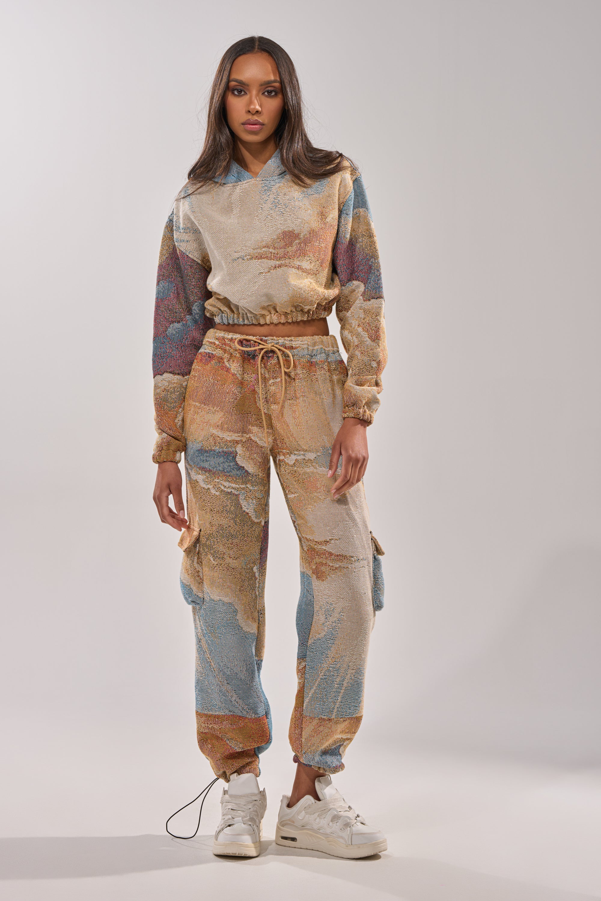 A woman with long, straight hair and a neutral expression faces forward in the INTO THE WILD WIDE LEG JOGGER and a matching hoodie with a colorful abstract print, paired with white sneakers against a plain light background. 0