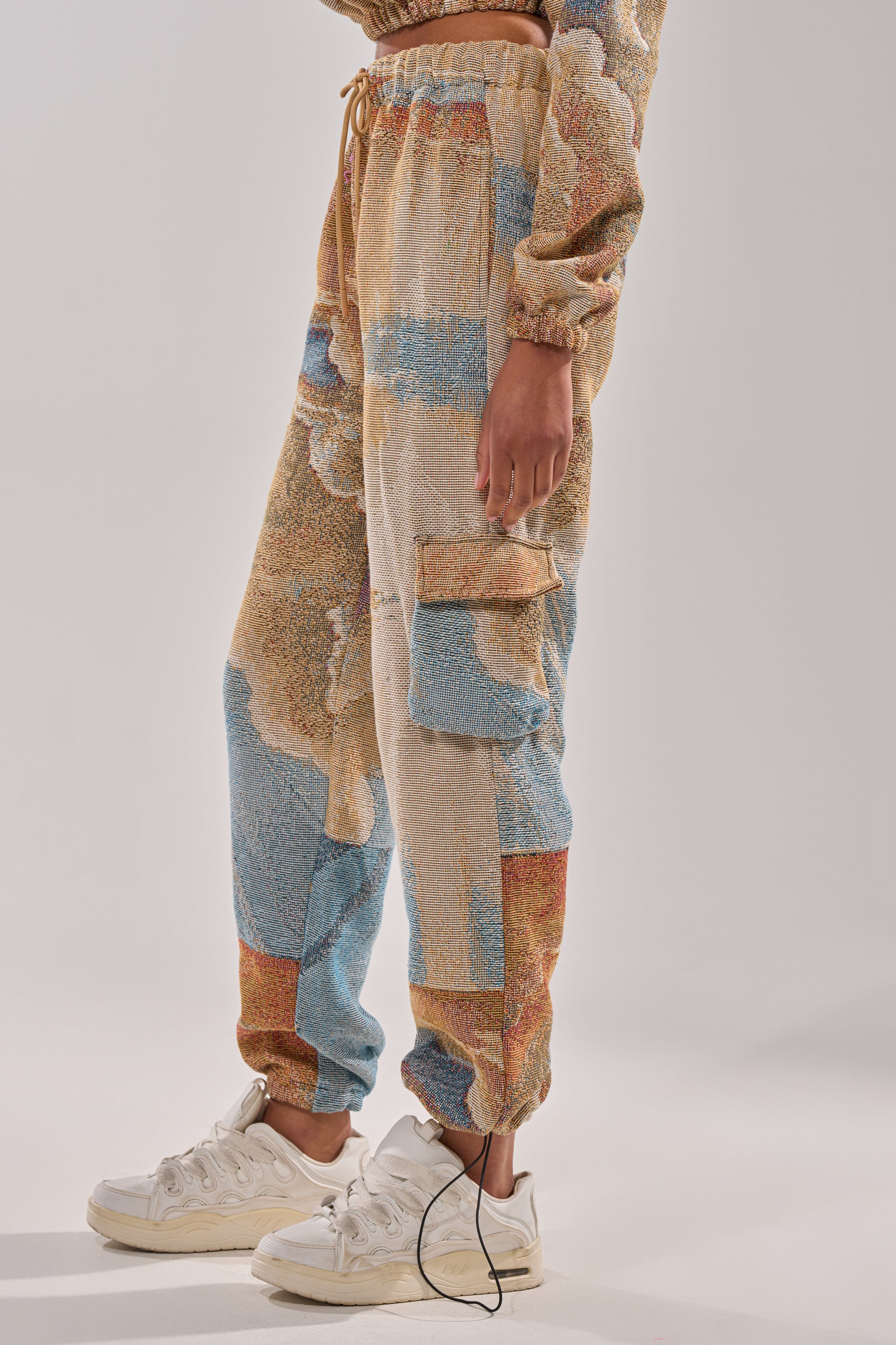 A person models the INTO THE WILD WIDE LEG JOGGER featuring a landscape pattern in beige, blue, and peach tones, paired with white sneakers. Their hand rests by their side against a plain light gray background. 2