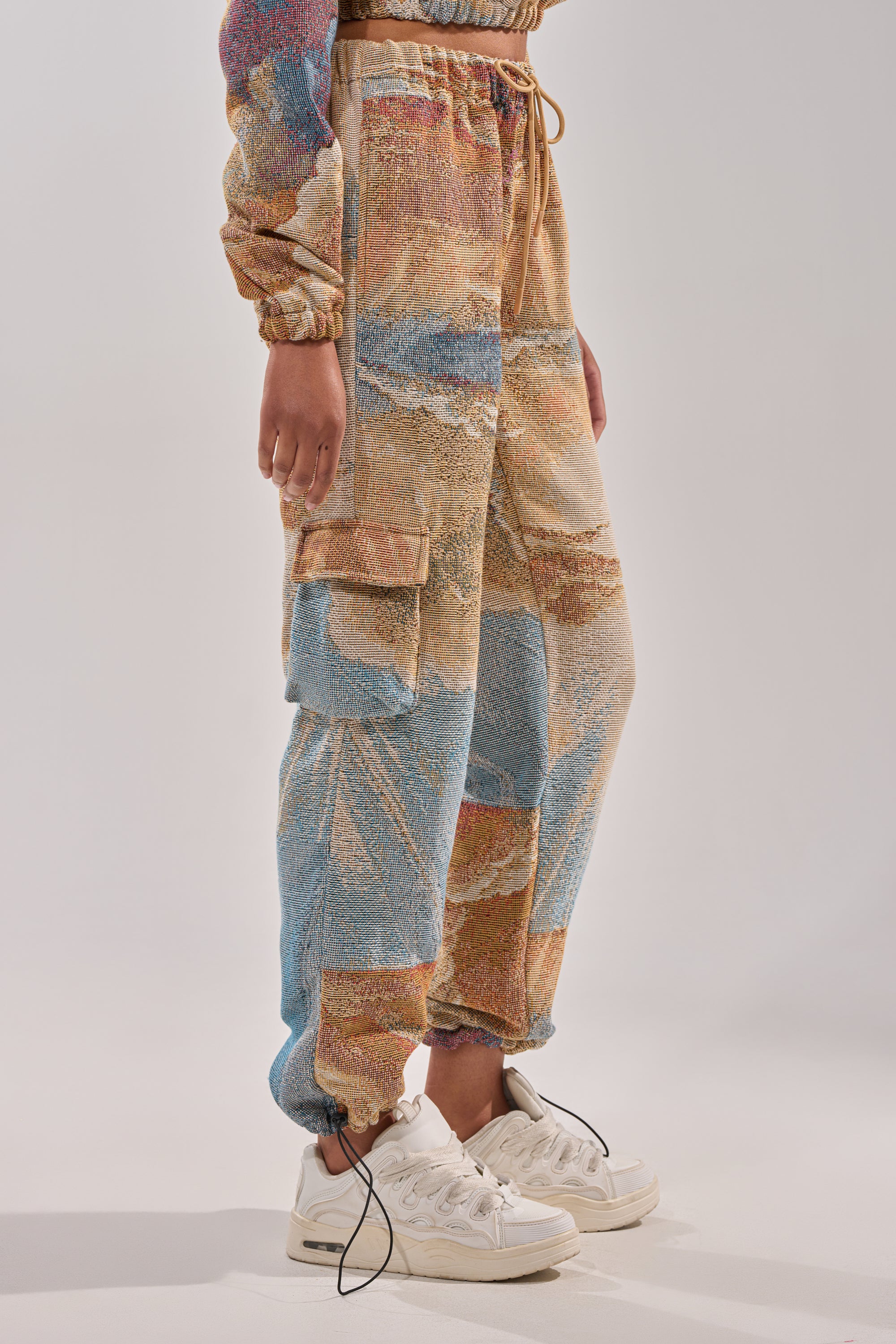 A person models the INTO THE WILD WIDE LEG JOGGER—streetwear pants with a multicolored abstract pattern, cargo pockets, and elastic cuffs—paired with white sneakers. The photo captures the waist down against a plain background. 3