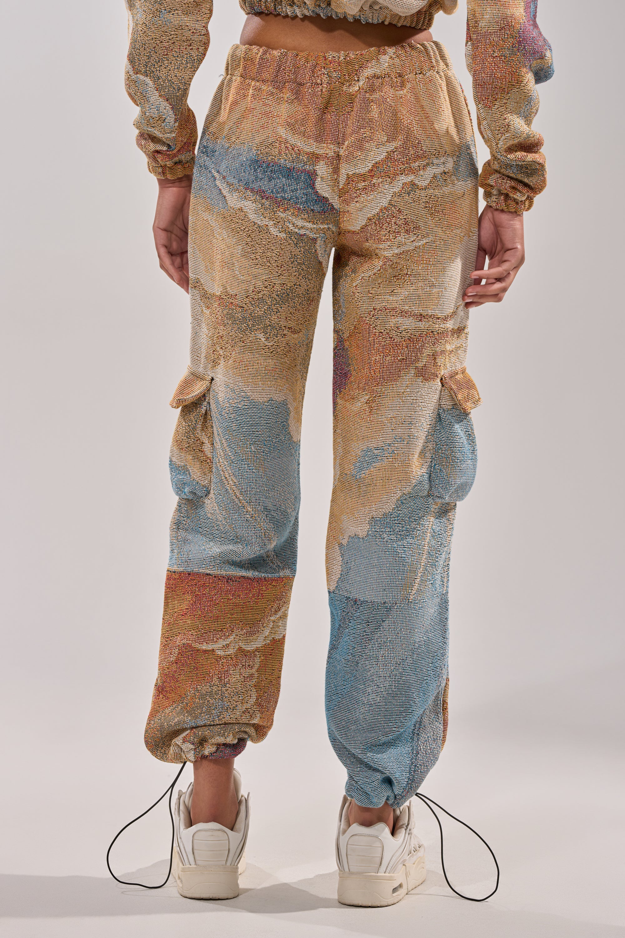 A person wears the INTO THE WILD WIDE LEG JOGGER, featuring a colorful, abstract pattern in beige, blue, and orange, paired with white sneakers. Photo taken from behind highlights its relaxed fit and cargo pockets for a fresh streetwear vibe. 4