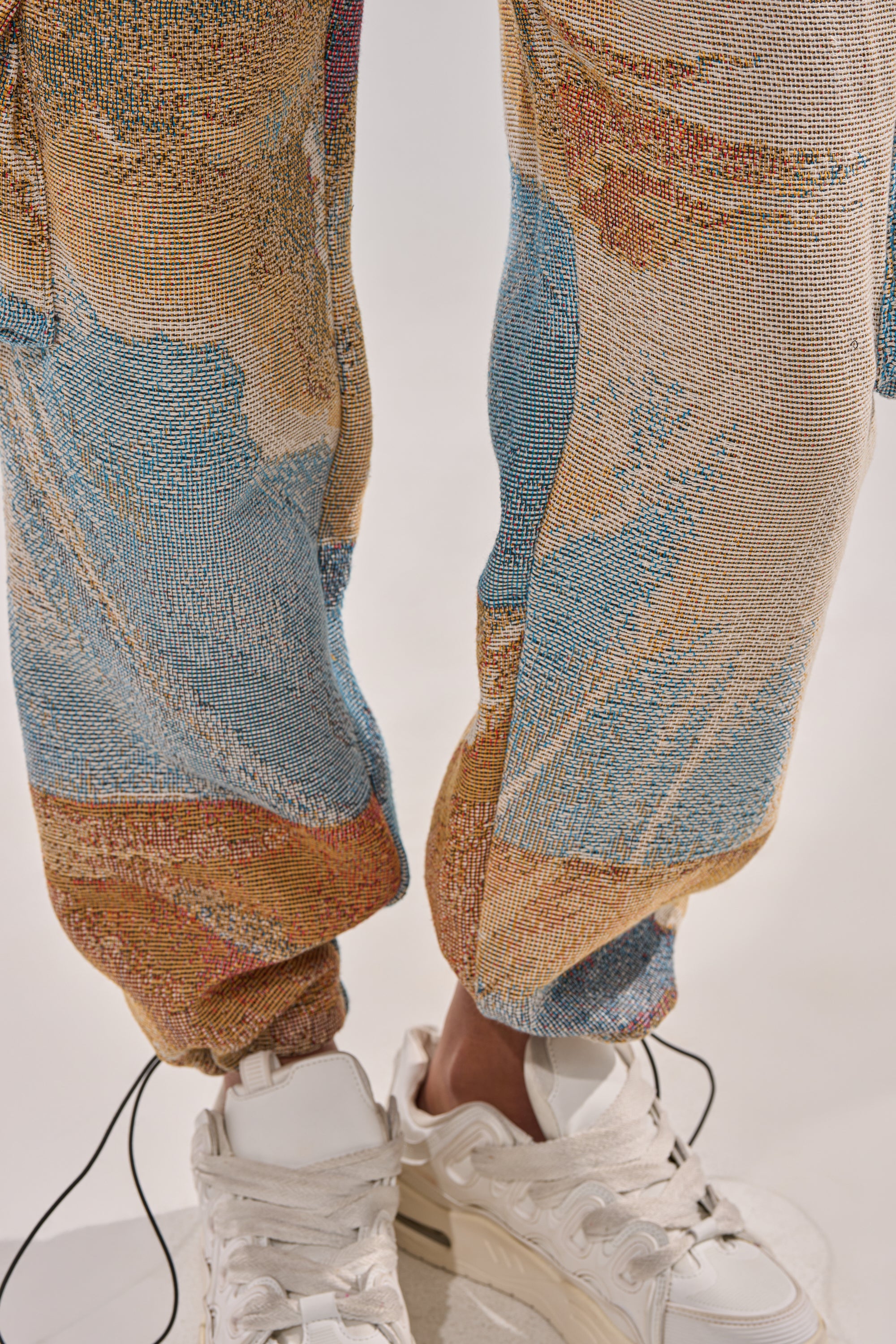 Close-up of a person wearing the INTO THE WILD WIDE LEG JOGGER, featuring an abstract blue, brown, and beige pattern. The joggers are gathered at the ankles and paired with chunky white sneakers against a plain light background. 7