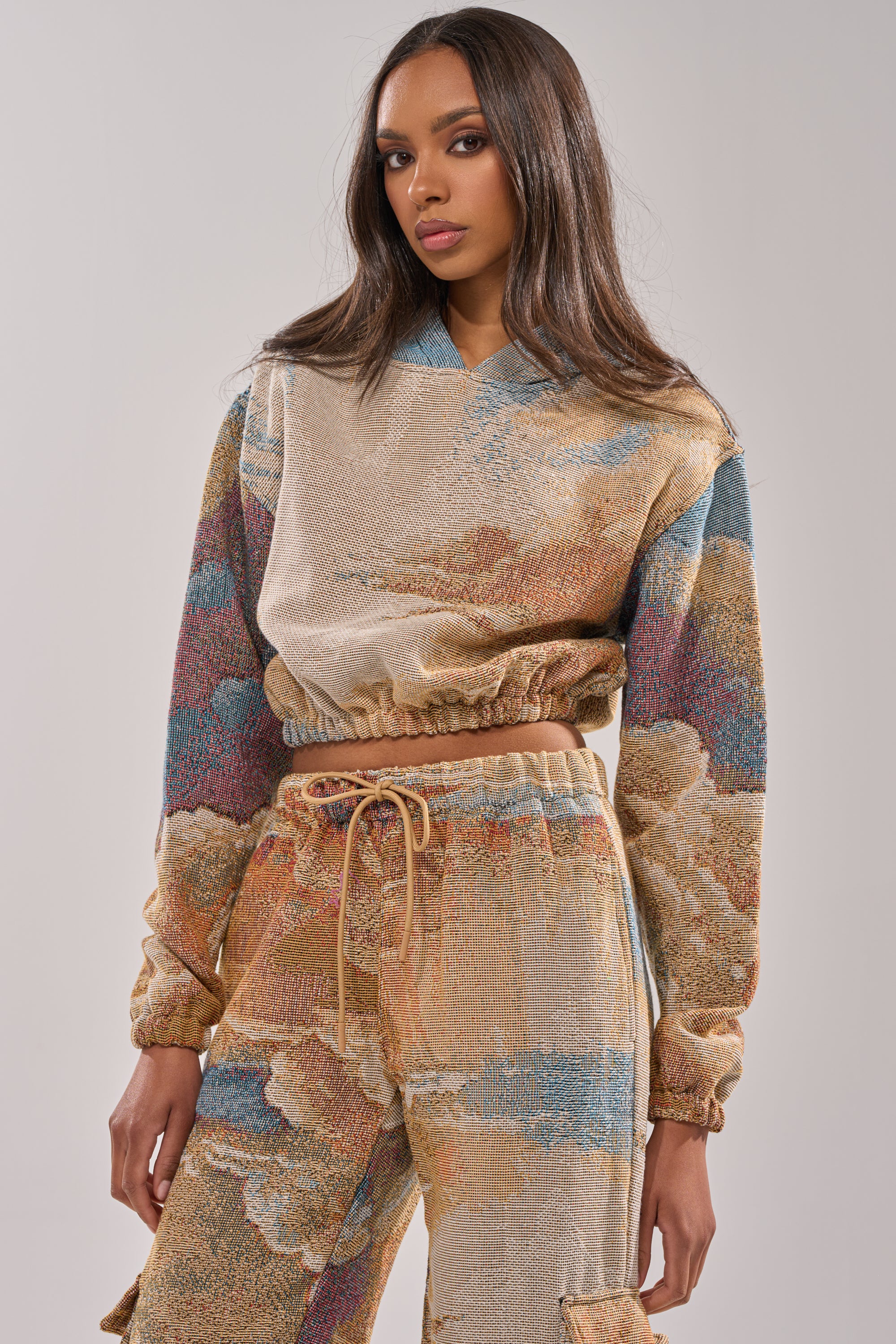A woman with long brown hair wears the INTO THE WILD SWEATSHIRT—a beige and multicolored tapestry cropped hoodie paired with matching drawstring pants—channeling vintage streetwear against a plain light background. 0