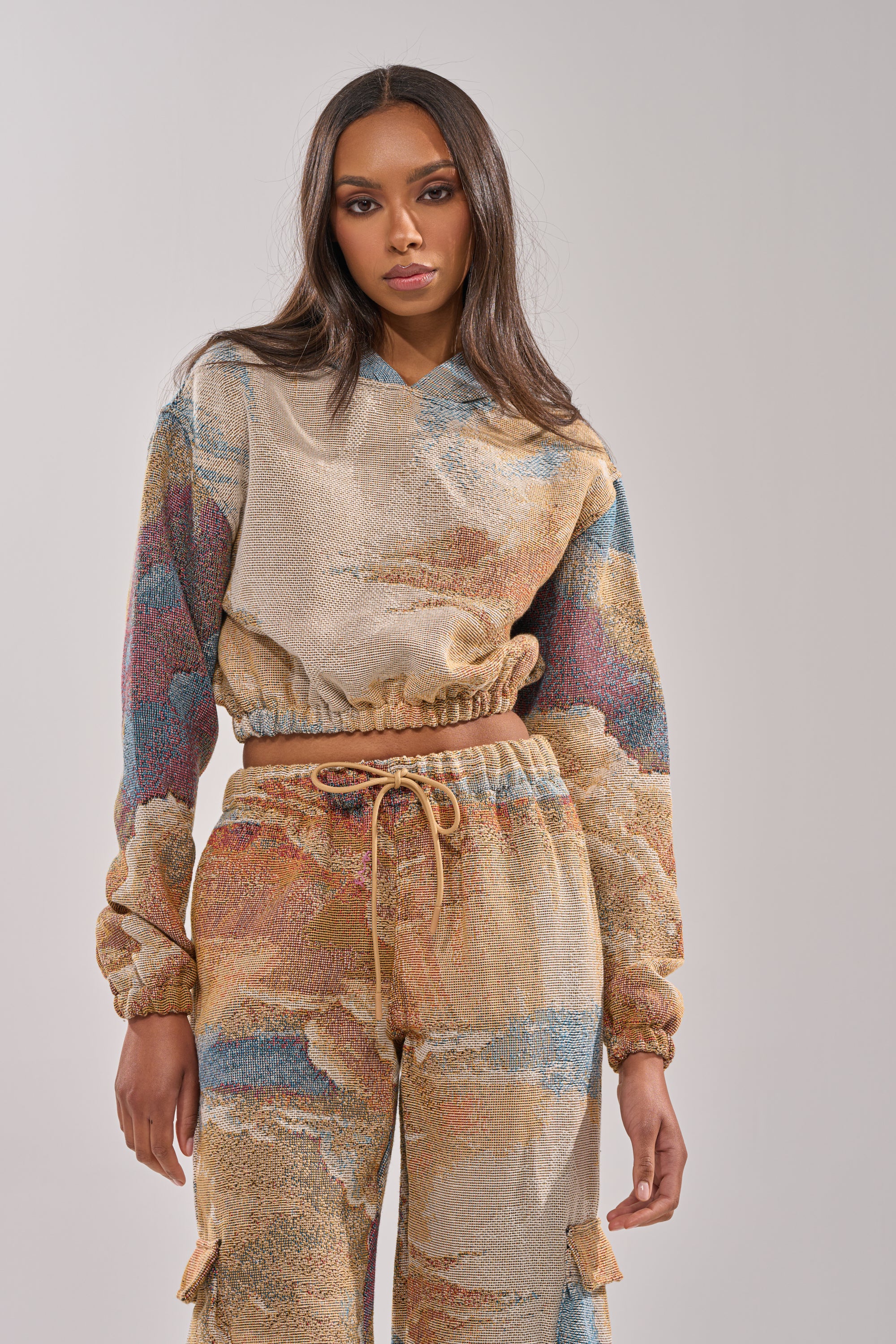 A woman wears the INTO THE WILD SWEATSHIRT, a tapestry cropped hoodie with an abstract, multicolored pattern, paired with matching high-waisted pants against a plain light background. 1