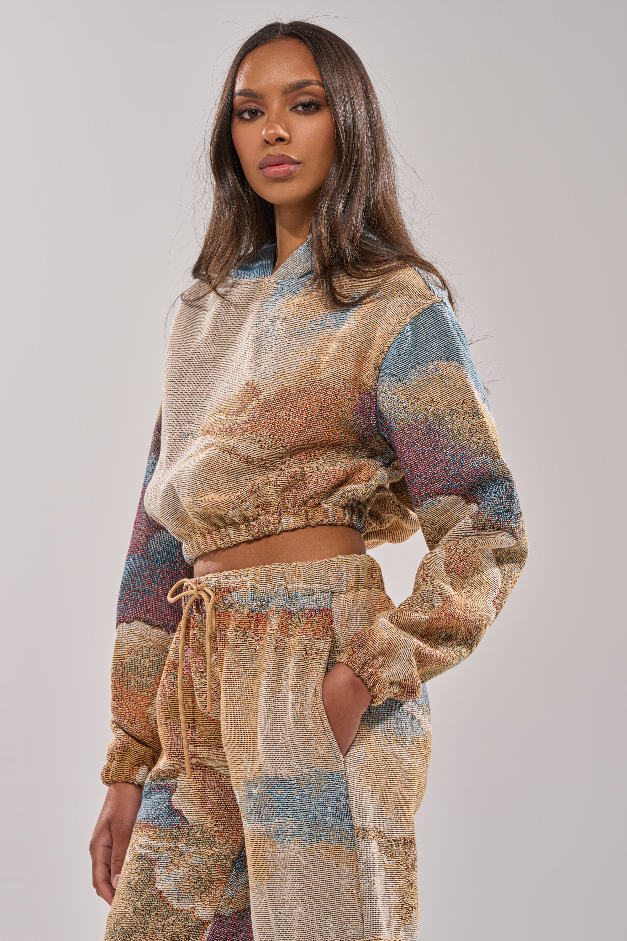 A woman with long brown hair wears the INTO THE WILD SWEATSHIRT, a tapestry cropped hoodie with earth-toned and blue abstract patterns, paired with drawstring pants for a vintage streetwear look against a plain light background. 2