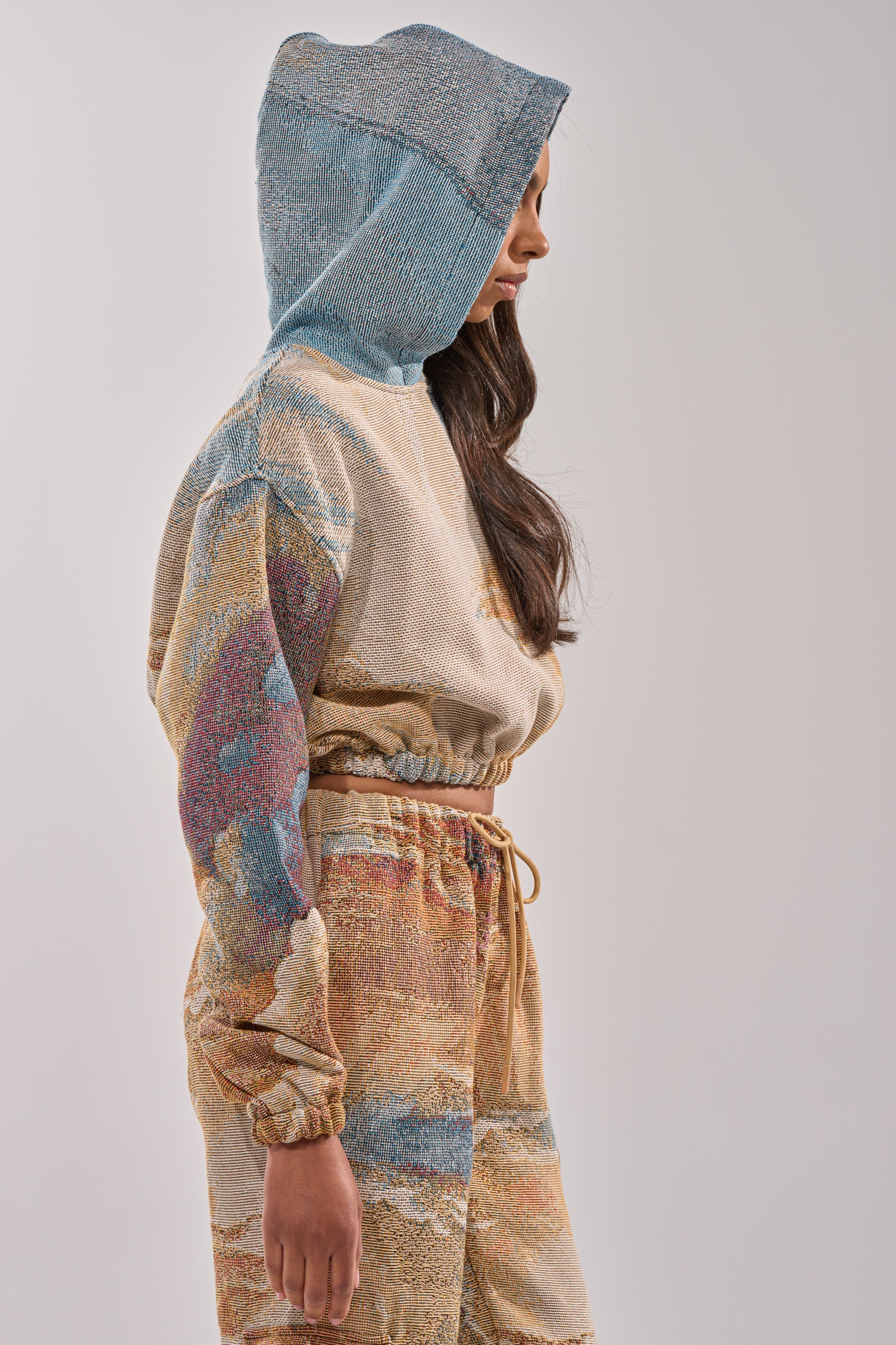 A person with long dark hair wears the INTO THE WILD SWEATSHIRT and matching pants in muted pastel blue, beige, and gold with an abstract vintage streetwear pattern, standing sideways against a plain light background. 3