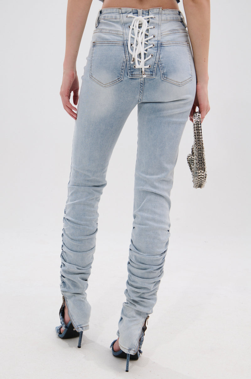 A person wearing the LACE IT UP SKINNY DENIM PANT, featuring a light-wash, high-rise fit with lace-up detail on the back and ruched legs, holds a silver chain purse and is photographed from behind against a plain background. 0