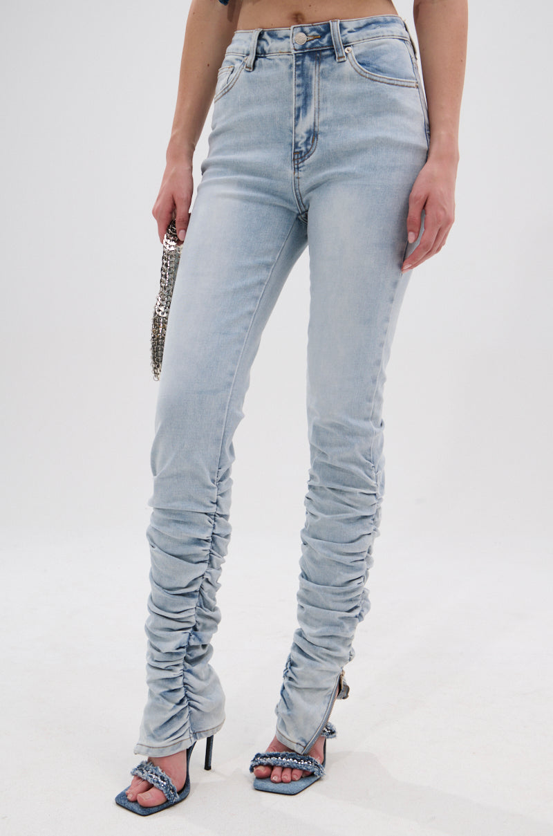 A person wears the LACE IT UP SKINNY DENIM PANT—light-wash, high-waisted jeans with corset lace-up details—styled with open-toe chain sandals and a metallic chain purse against a plain white background. 1