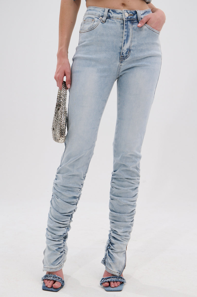 A woman models the LACE IT UP SKINNY DENIM PANT, a high-waisted, light-wash ruched skinny jean, paired with blue open-toe heels and a small silver bag, standing against a white background. 2