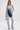 A woman stands confidently in the LACE IT UP SKINNY DENIM PANT, paired with a halter-style denim top featuring a dark diagonal crossbody detail. She carries a patterned handbag and completes her look with chic high-heeled sandals.
