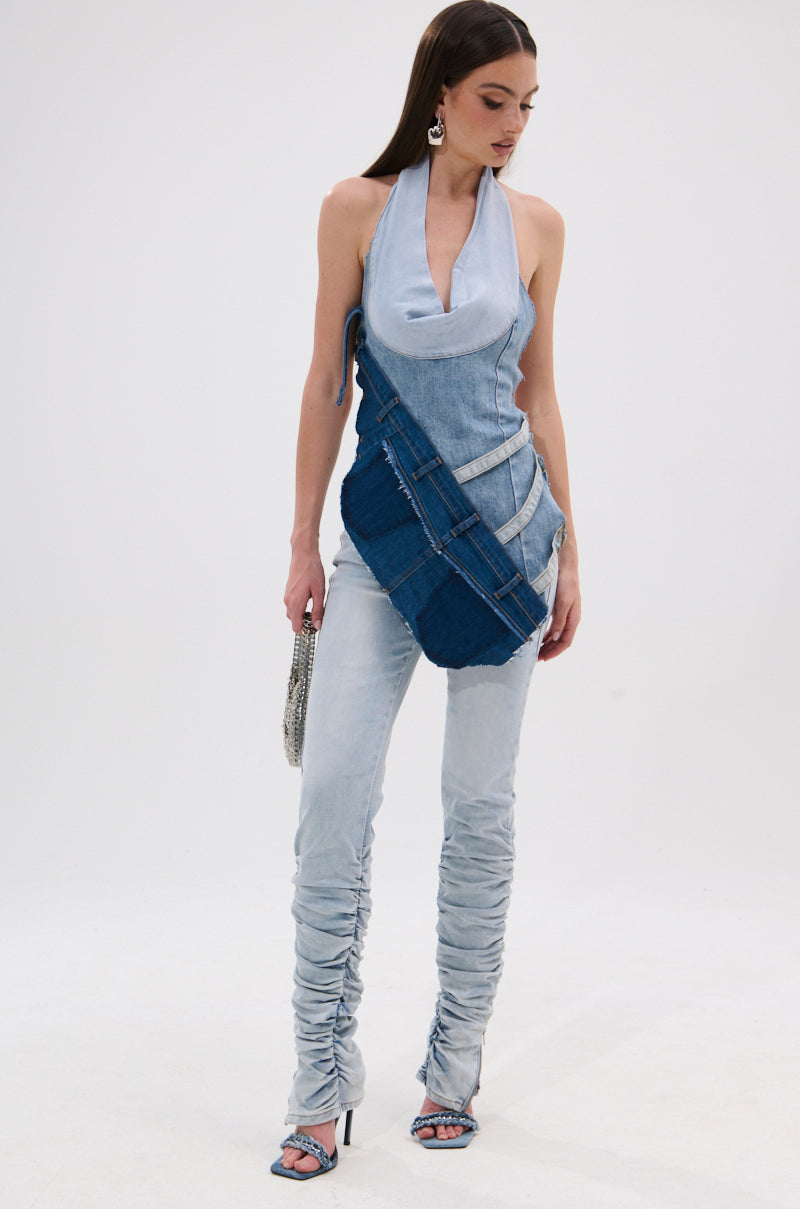 A woman stands against a white background wearing the LACE IT UP SKINNY DENIM PANT with ruched legs, a light denim halter top, dark denim crossbody sash, silver earrings, high-heeled sandals, and holding a small purse. 5