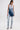 A woman stands against a white background wearing the LACE IT UP SKINNY DENIM PANT with ruched legs, a light denim halter top, dark denim crossbody sash, silver earrings, high-heeled sandals, and holding a small purse.