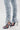 A close-up of a person wearing the LACE IT UP SKINNY DENIM PANT in light blue, high-rise fit, skinny style with zippered ankles, paired with high-heeled open-toe sandals with textured straps against a plain, light background.