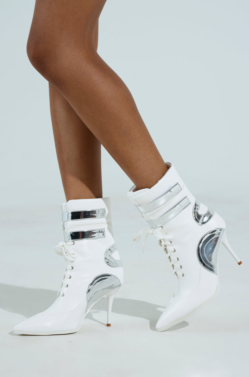 Someone wearing the AZALEA WANG LACIE SILVER SPORTY BOOTIE, featuring white vegan leather, stiletto heels, metallic details, and lace-up fronts for a sleek and futuristic look against a plain light background.