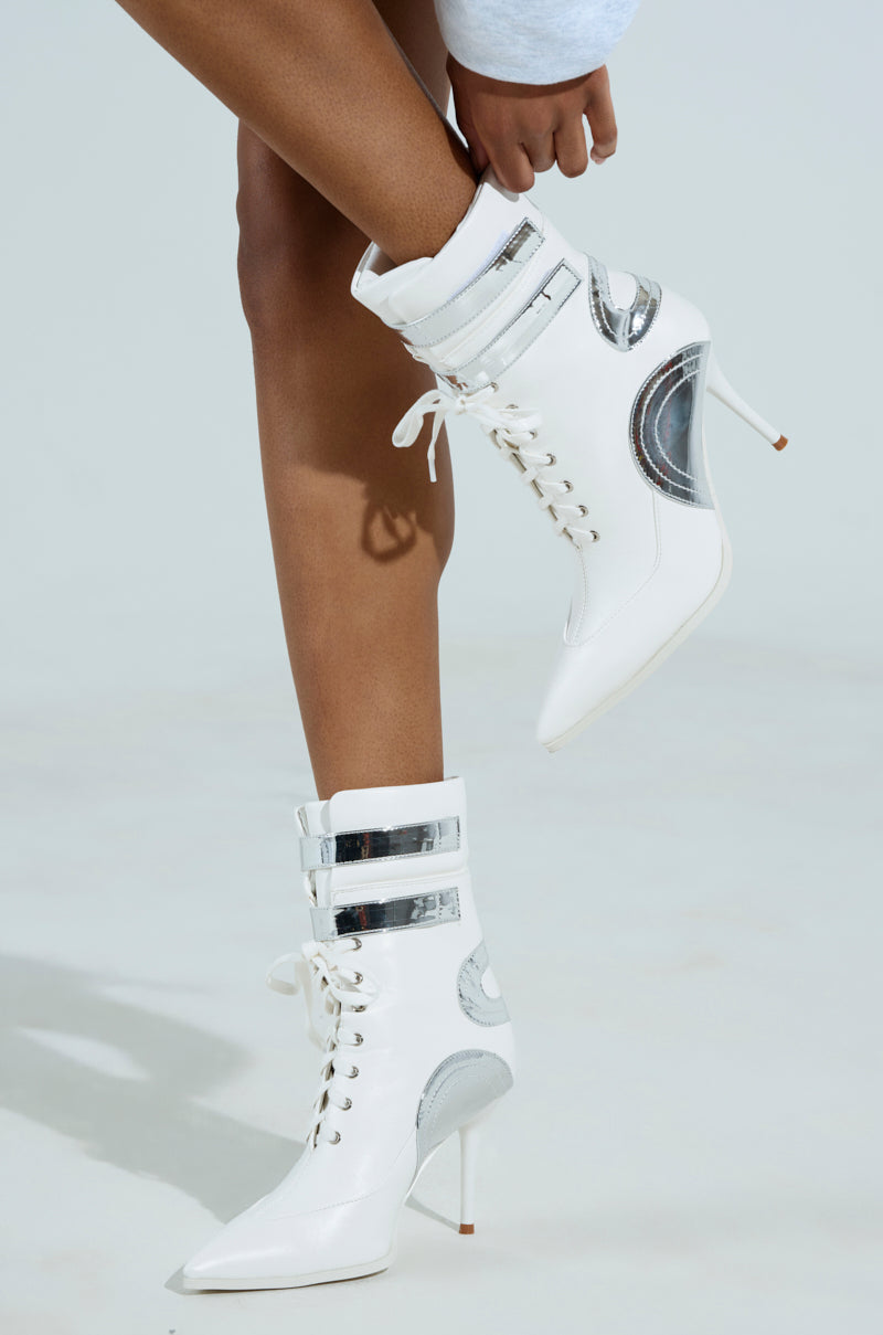 A person wears the chic AZALEA WANG LACIE SILVER SPORTY BOOTIE, featuring stylish silver accents and laces. The eye-catching metallic details and sleek stiletto heel stand out against a plain light background, highlighting the elegance of their pose.