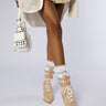 A person stands modeling a stylish outfit, featuring a beige mini skirt, a matching jacket with perforated detailing, AZALEA WANG LACIE SUEDE BOOTIE paired with white lace socks, and a white handbag adorned with metal studs. The background is plain and light-colored.