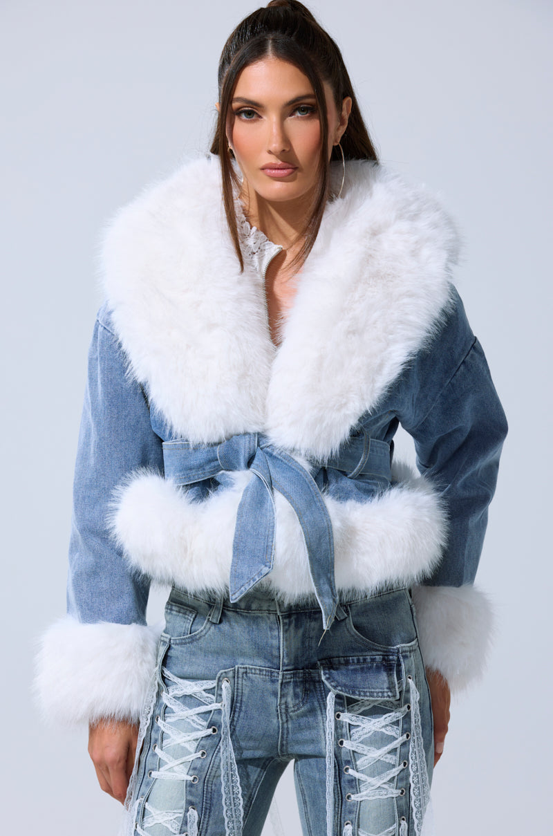 A woman models the LAINEY FUR TRIM DENIM MOTO—a cropped denim jacket with a bold white faux fur collar and cuffs—paired with denim lace-up pants with white ribbons, standing confidently against a plain light background.