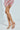 A person stands with crossed legs, wearing AZALEA WANG LAMIUM ROSE GOLD LEAF SANDALS with stiletto heels, elegantly paired with a colorful fringed skirt against a light, neutral backdrop.