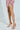 A person elegantly showcases their legs from the knees down, wearing chic AZALEA WANG Lamium Rose Gold Leaf Sandals with stiletto heels, against a light background. They are dressed in a colorful fringe-textured skirt or dress.