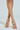 Crossed legs wear AZALEA WANG LAMIUM ROSE GOLD LEAF SANDALS, showcasing their open toes and stiletto heels made from vegan leather. The metallic leaf designs enhance the skin tone against a soft blue background, highlighting the allure of these elegant shoes.