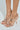 A pair of feet wear the AZALEA WANG LAMIUM ROSE GOLD LEAF SANDALS, featuring stiletto heels, metallic leaf details, and transparent straps with open toes in vegan leather. The light background enhances their elegance.